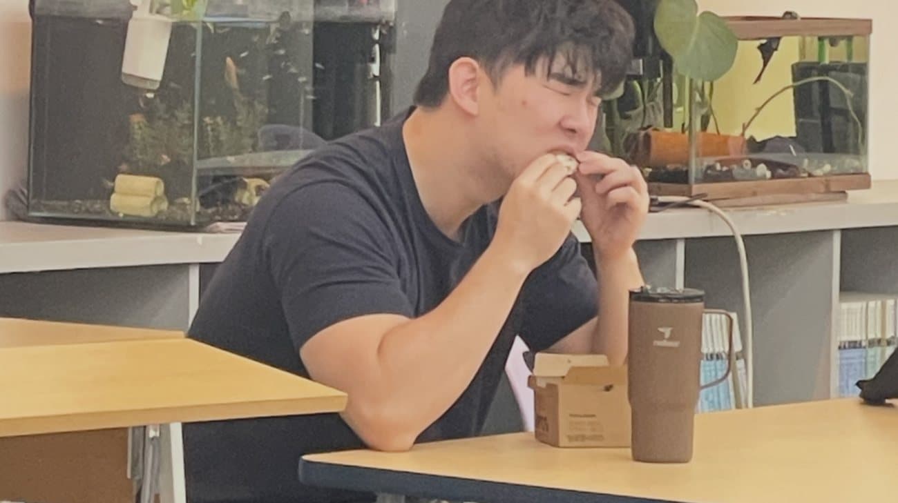 A student at a classroom desk, eyes squeezed shut, biting into a snack