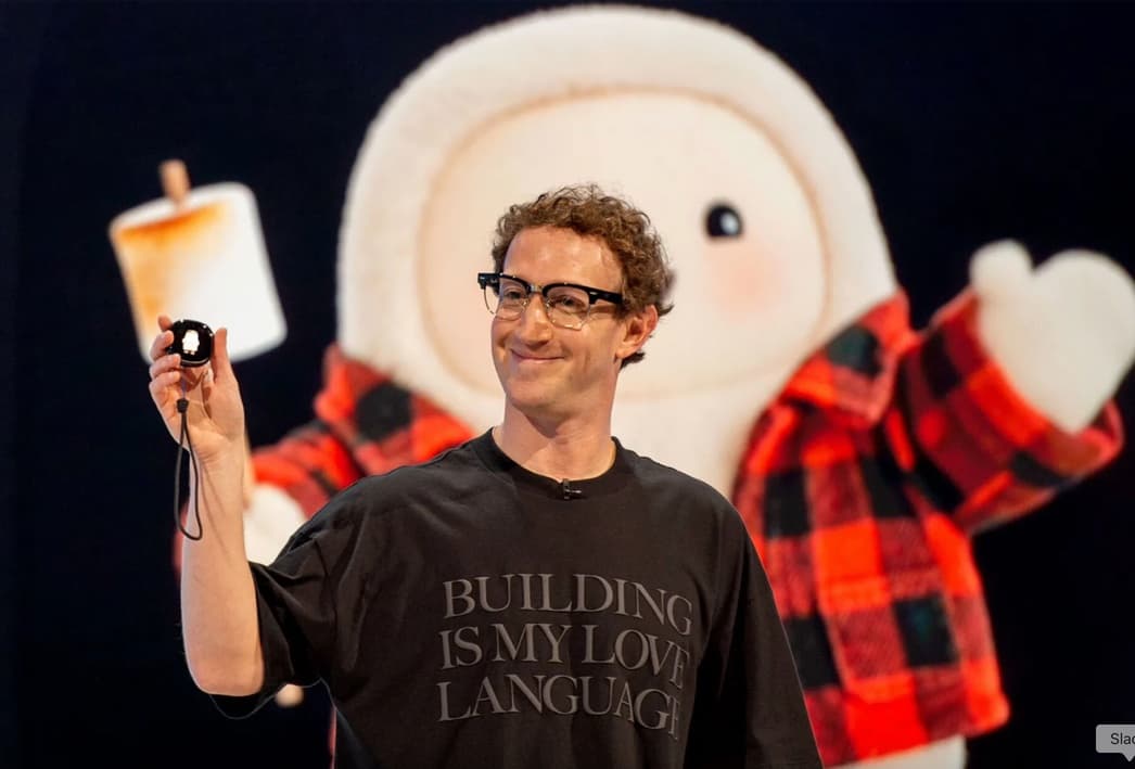Mark Zuckerberg on stage holding up a small device, a giant plush mascot in a plaid shirt behind him