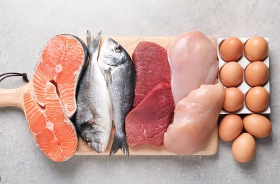 Raw salmon, whole fish, beef, chicken breast and a tray of eggs laid out on a wooden board