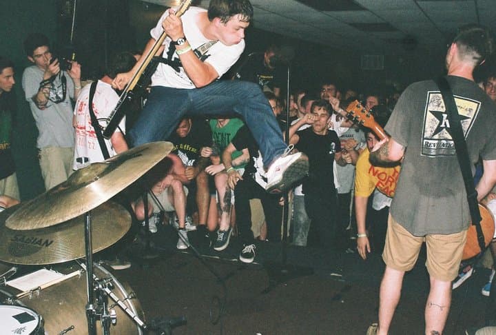 A singer leaping above a packed basement crowd at a hardcore show, drum kit in the foreground