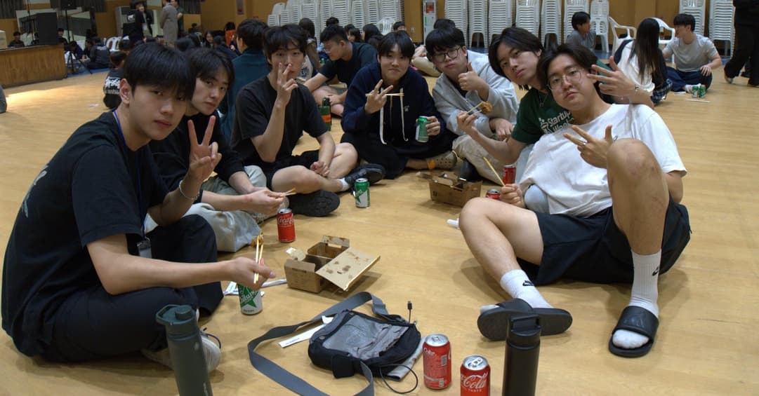 Students sitting in a circle on a gym floor with snacks and cans of soda, flashing peace signs at the camera